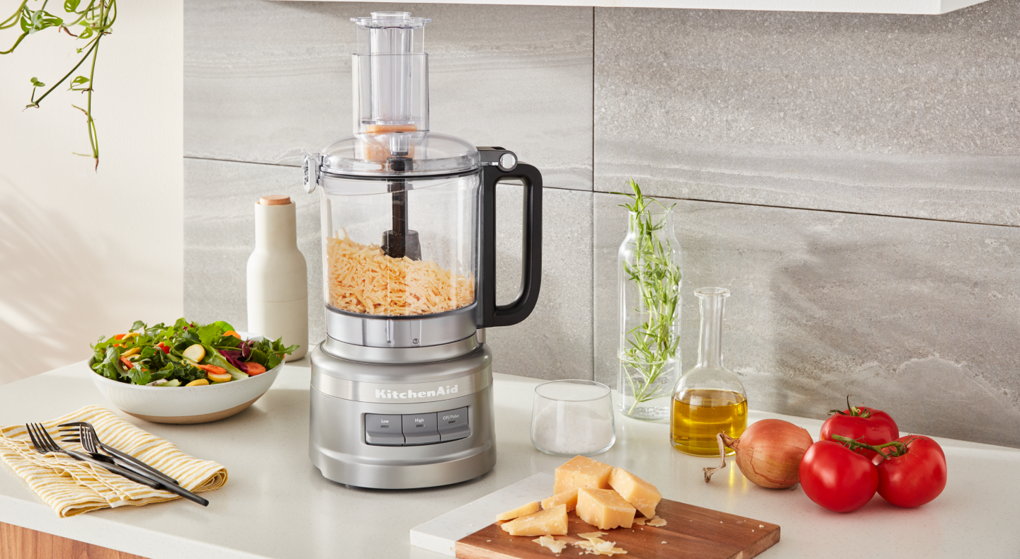 Shredded cheese sitting in work bowl of silver food processor on the counter with tomatoes, salad and other ingredients 