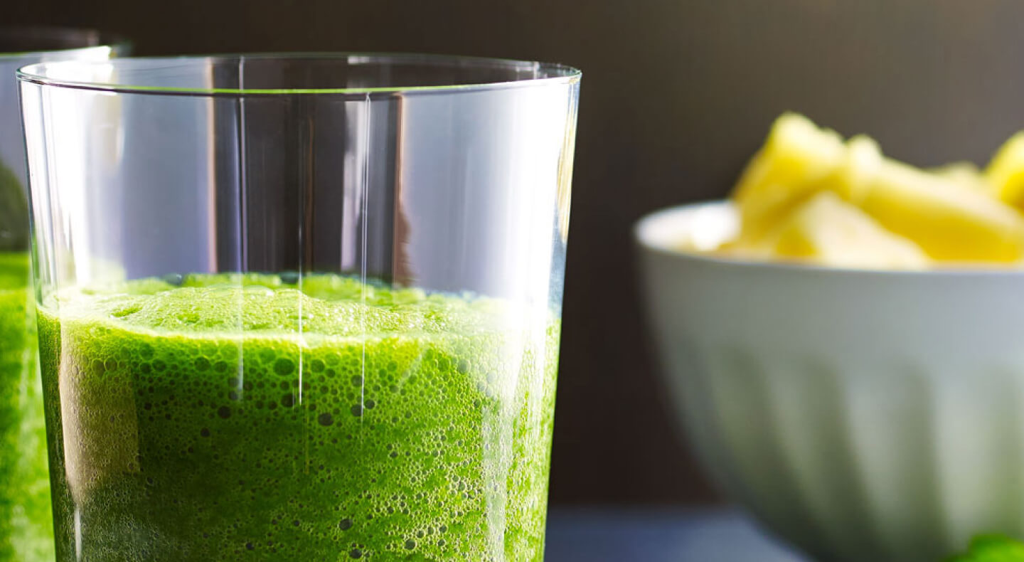 Green juice in a glass next to a bowl of sliced pineapples