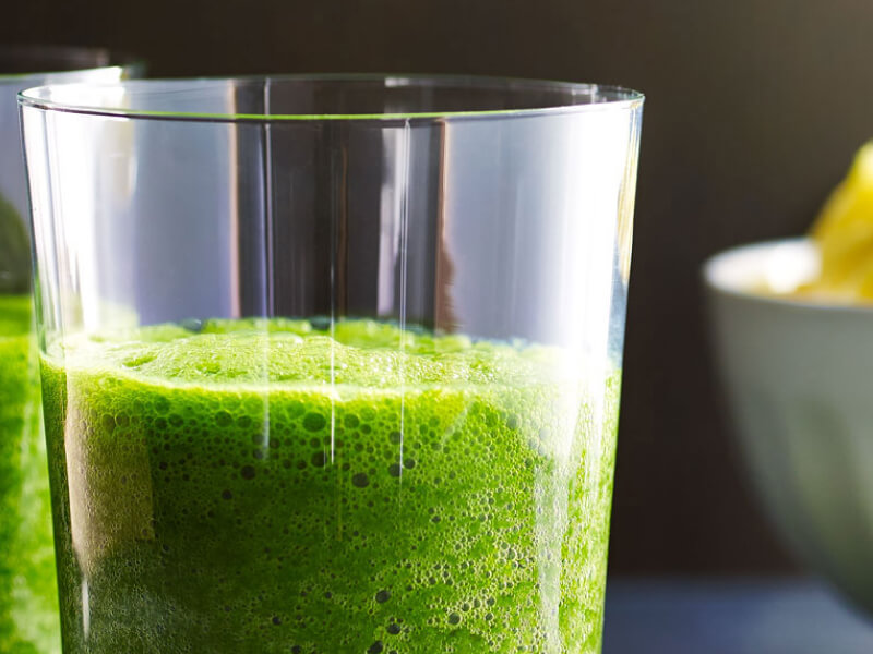 Green juice in a glass next to a bowl of sliced pineapples