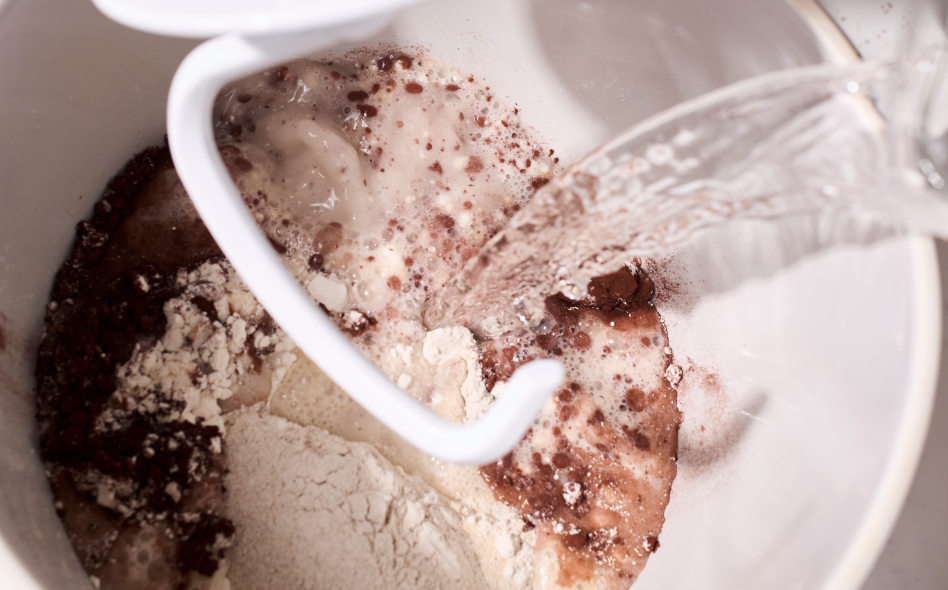 Close up of water being poured into mixing bowl with dry ingredients and dough hook