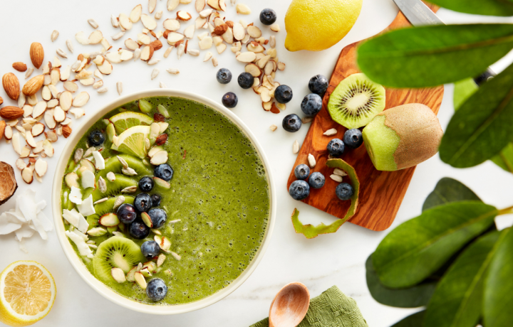 Overhead shot of the perfect green smoothie bowl surrounded by blueberries, seeds, sliced kiwi, lemon and almonds
