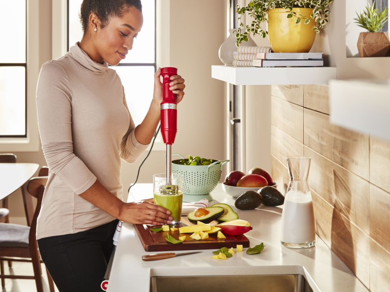 Red KitchenAid® immersion blender pureeing food in a glass next to various ingredients