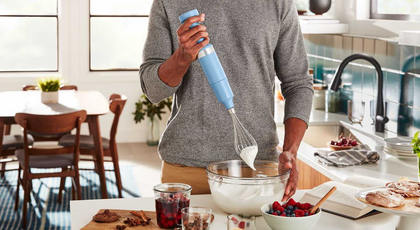 A man using a KitchenAid® hand blender to whip egg white peaks in a modern kitchen. 