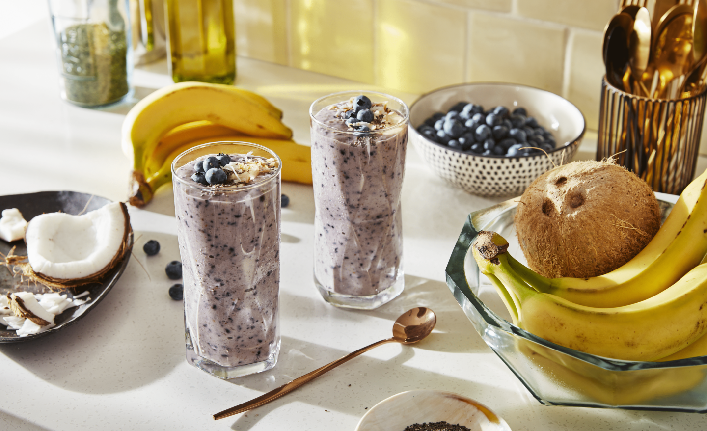 Two blueberry smoothies on countertop with bananas and coconut ingredients