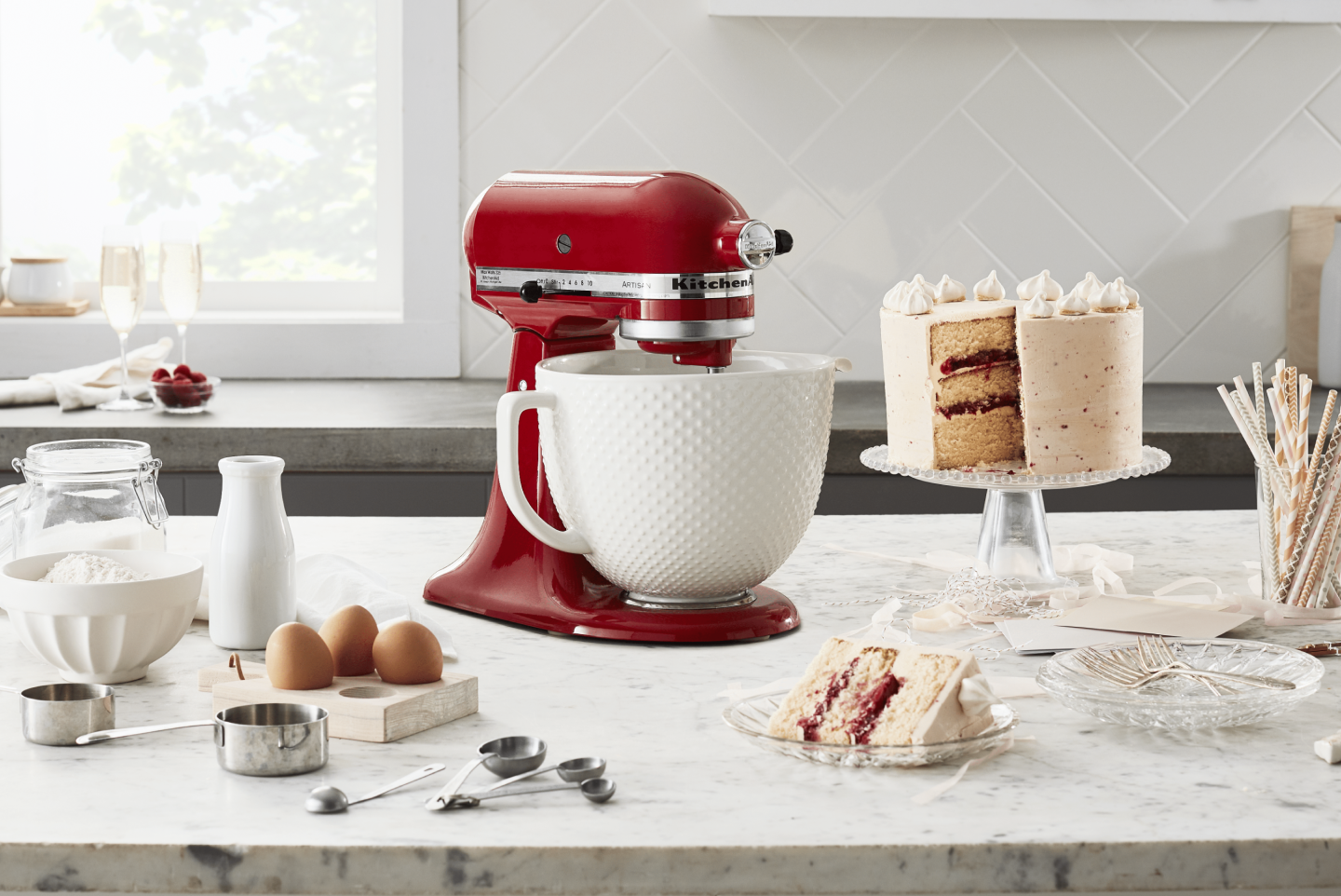 Red KitchenAid® Stand Mixer on a counter next to a three layer white frosted cake, milk, eggs, measuring cups and flour 
