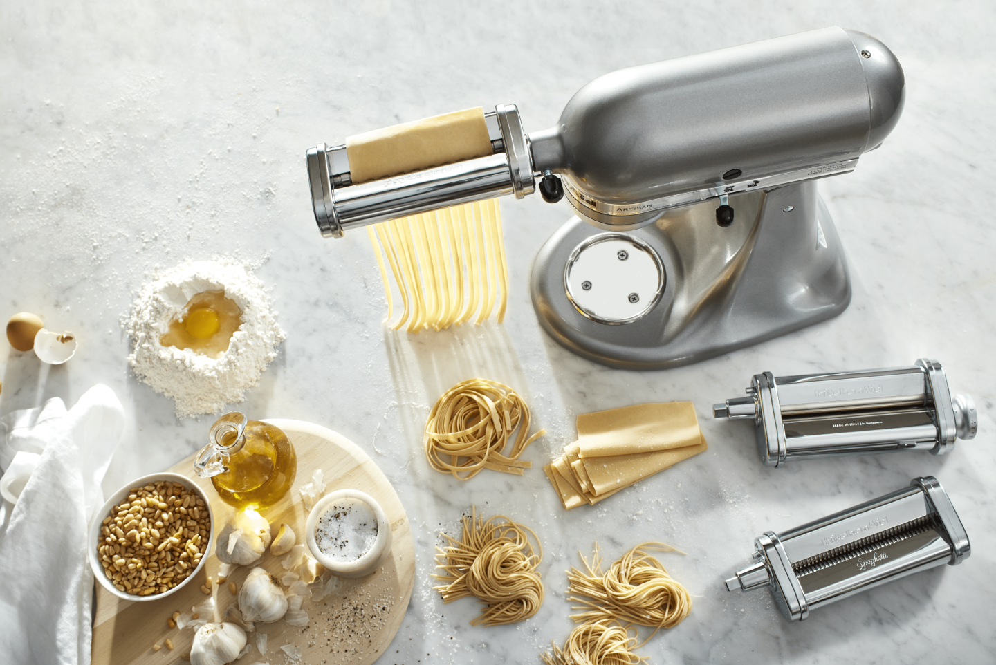 Silver KitchenAid® Stand Mixer with pasta-making attachments surrounded by mounds of fresh pasta and pasta ingredients 
