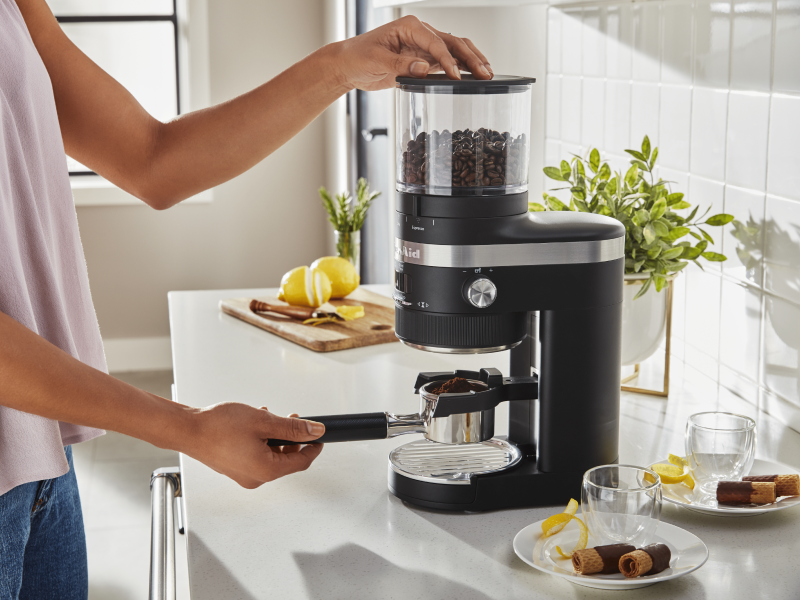 A woman using a black KitchenAid® Burr Coffee Grinder to process coffee beans