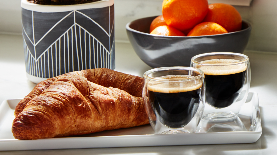 Croissants and cups of espresso on a tray in front of a bowl of oranges.