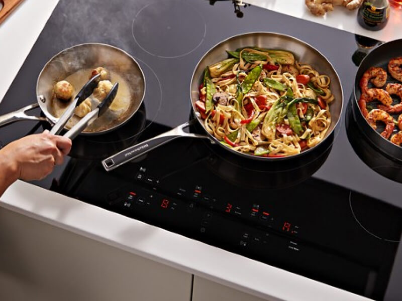 A person cooking pasta and shrimp on a black KitchenAid® electric induction cooktop