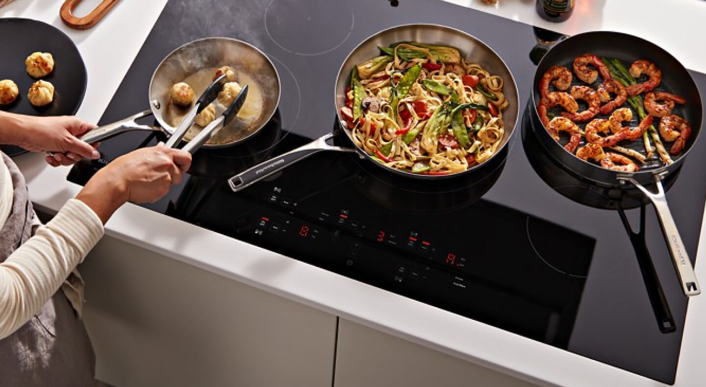 A person cooking pasta and shrimp on a black KitchenAid® electric induction cooktop