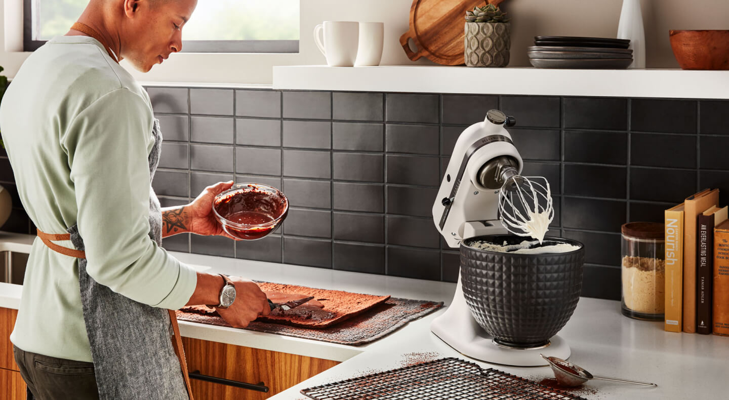 A person with a bowl of melted chocolate standing next to a KitchenAid® stand mixer with a wire whisk accessory and whipped egg whites.
