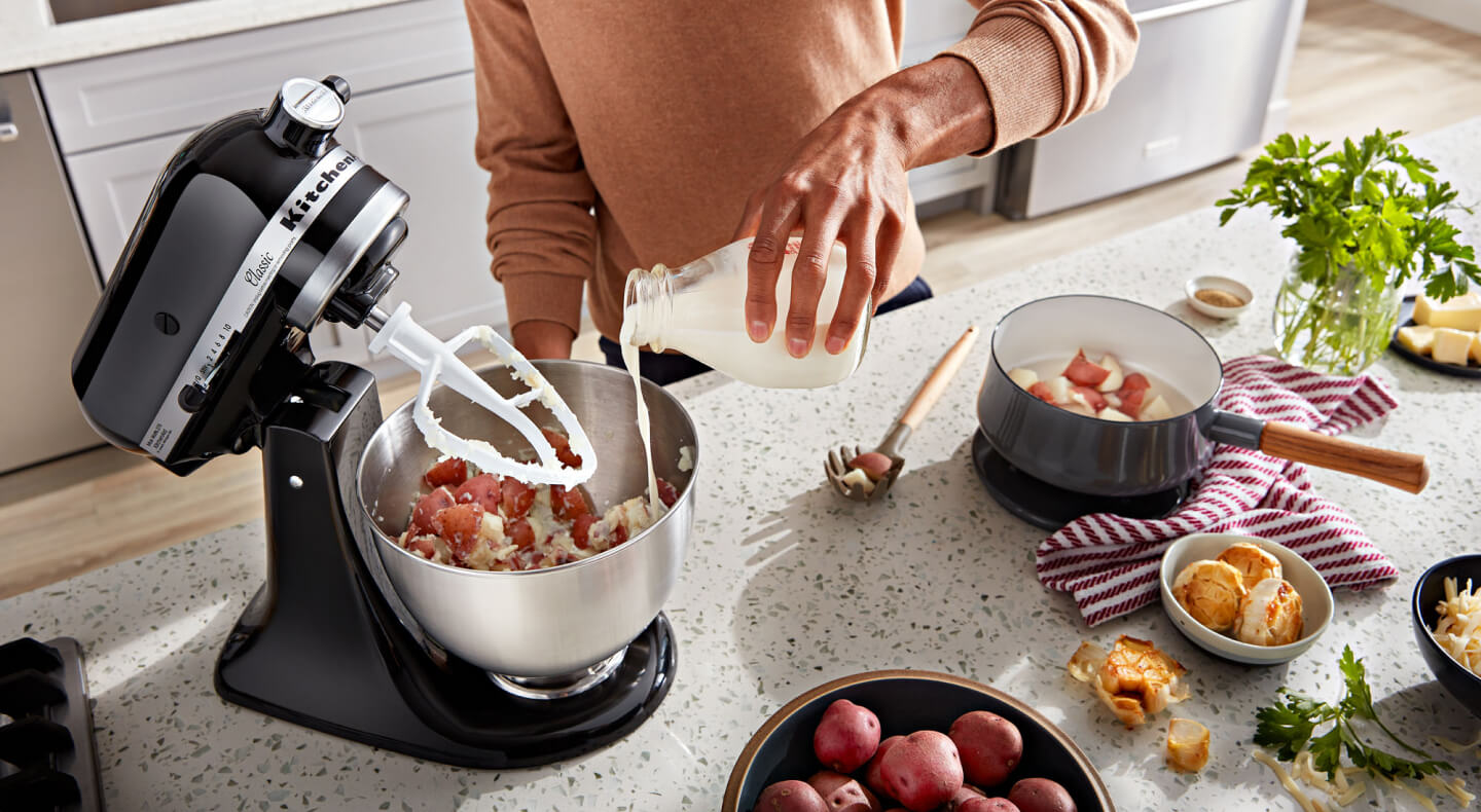 A person pouring milk into a KitchenAid® stand mixer bowl with ingredients and a flat beater accessory on the counter of a modern kitchen.