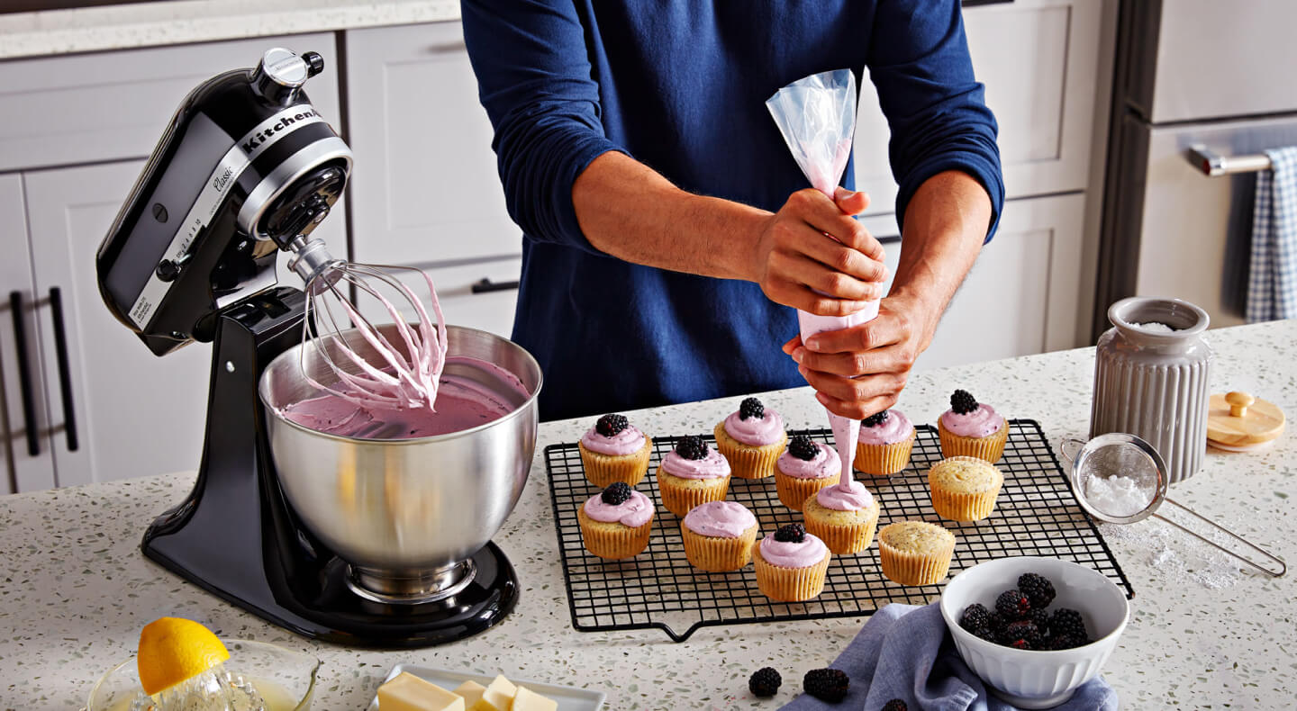 A person frosting cupcakes on the counter of a modern kitchen next to a KitchenAid® stand mixer with a wire whisk/whip accessory.
