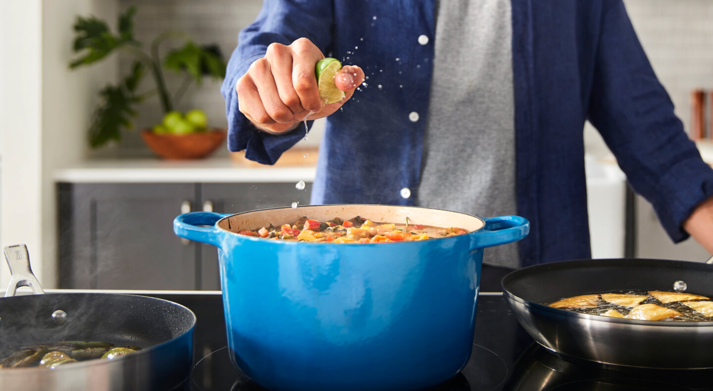 A person squeezing a lime over a rich stew cooking in a blue Dutch oven