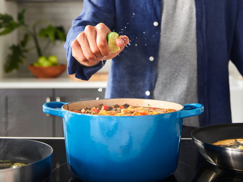 A person squeezing a lime over a rich stew cooking in a blue Dutch oven