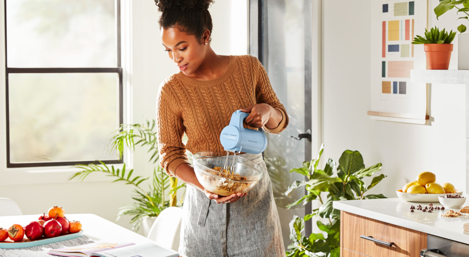 A woman reading a cookbook and holding a cordless hand mixer and a bowl