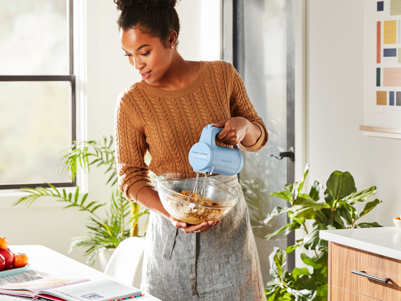 A woman reading a cookbook and holding a cordless hand mixer and a bowl