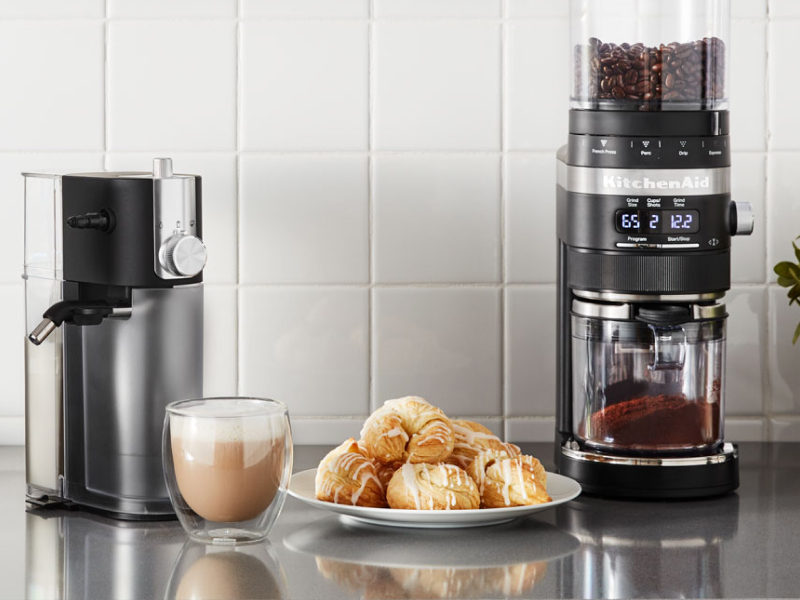 A cup of whipped coffee on a stainless steel countertop with baked goods and appliances