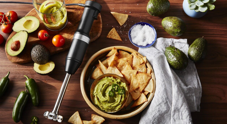  Immersion blender laying beside bowl of guacamole and chips surrounded by guacamole ingredients