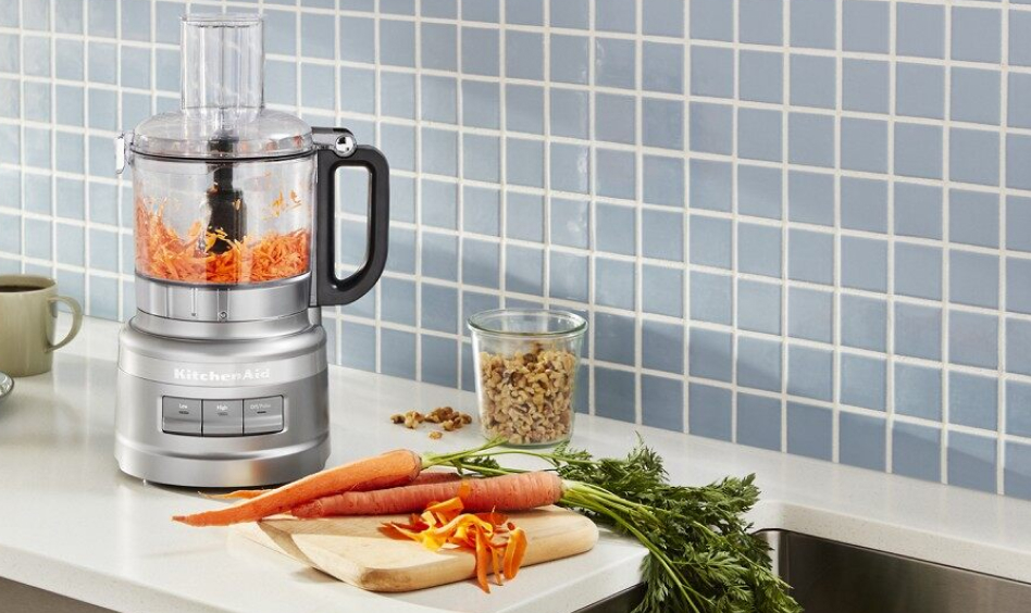 Silver KitchenAid® Food Processor on counter with carrots and blue backdrop
