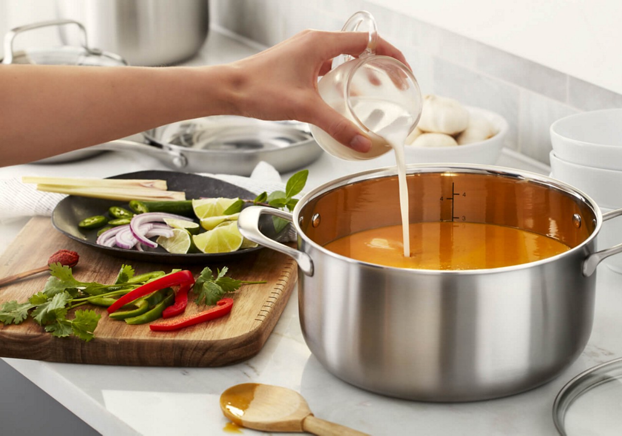 Pouring cream into pan of soup sitting next to cutting board with herbs, peppers and lime wedges