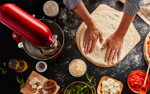 Person flattening out pizza dough on countertop with KitchenAid® stand mixer and pizza ingredients